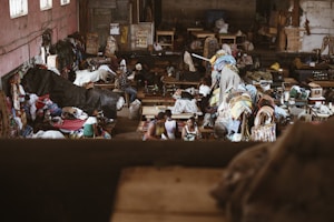 The image depicts a bustling indoor market or workshop filled with textiles and people. Various colorful fabrics and clothes are scattered throughout the room, with sewing machines visible in the background. People are engaged in activities related to the textile industry, possibly working or trading. The environment appears lively and somewhat chaotic, with a mixture of colors and textures.