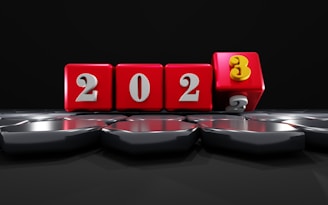 Red cubes with white numbers form the sequence 2023. The cube displaying the number 3 is tilted forward, replacing the number 2. The cubes are placed on a reflective surface with hexagonal patterns under a dark backdrop, creating a dynamic presentation of the new year.