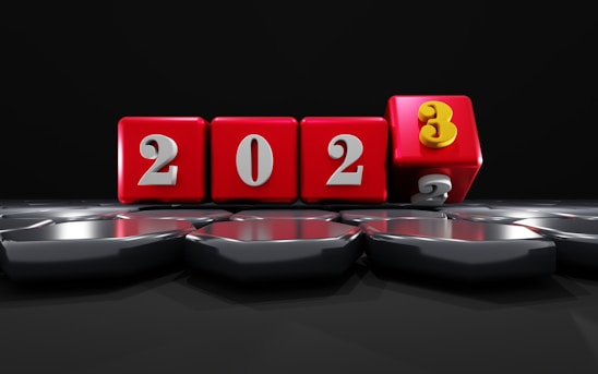 Red cubes with white numbers form the sequence 2023. The cube displaying the number 3 is tilted forward, replacing the number 2. The cubes are placed on a reflective surface with hexagonal patterns under a dark backdrop, creating a dynamic presentation of the new year.