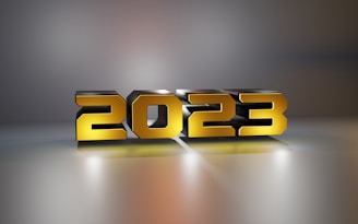Large three-dimensional numbers displaying 2023 are designed with a metallic, reflective, yellow and silver finish. The numbers are illuminated from below, casting a subtle glow on the surface and creating soft shadows on a gradient gray background.