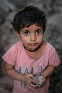 A young child with large expressive eyes is holding a stone or clump of mud in their hands. The child is wearing a pink shirt with the word 'HAPPY' partially visible. Their face and hands appear to be slightly dirty, suggesting they've been playing outdoors.