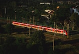 A red and white train travels along a railway track surrounded by dense green foliage. In the background, there are several buildings partially obscured by trees. The scene is captured from an elevated perspective, providing a view of the surrounding landscape and the expanse of track.