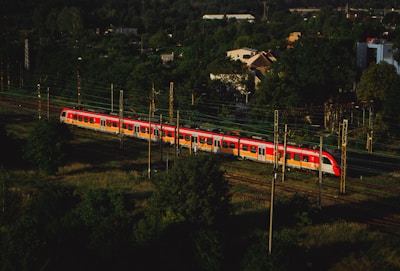 A red and white train travels along a railway track surrounded by dense green foliage. In the background, there are several buildings partially obscured by trees. The scene is captured from an elevated perspective, providing a view of the surrounding landscape and the expanse of track.