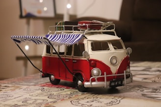 A detailed model of a vintage red and white VW camper van sits on a table. The van features a striped blue and white awning attached to its side and several items are secured to the roof. Soft lighting and a blurred background suggest an indoor setting.