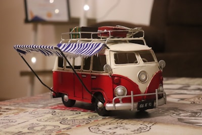 A detailed model of a vintage red and white VW camper van sits on a table. The van features a striped blue and white awning attached to its side and several items are secured to the roof. Soft lighting and a blurred background suggest an indoor setting.