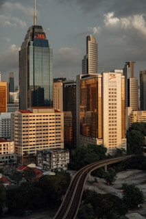 Tall skyscrapers dominate the cityscape, with glass facades reflecting the muted sunlight. A prominent building with a sharp spire bears the initials 'JKG'. In the foreground, a monorail track curves through the city, surrounded by lush trees and smaller structures. The sky is partly cloudy, adding to the urban ambiance.