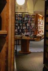 An indoor scene of a cozy bookstore with shelves filled with books. The store has wooden tables and shelves, and stacks of books arranged on them. The lighting is warm and inviting, and a small statue and a bright round light can be seen at the top center.