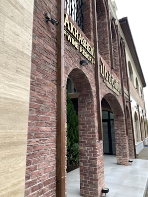 A building facade with red brick arches and large windows. The signage indicates a wine resort called 'Akhashen'. The architectural style is rustic with a modern touch, featuring lantern-style lights attached to the walls.