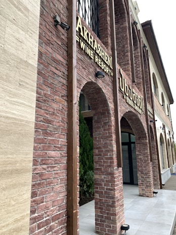 A building facade with red brick arches and large windows. The signage indicates a wine resort called 'Akhashen'. The architectural style is rustic with a modern touch, featuring lantern-style lights attached to the walls.