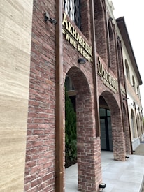 A building facade with red brick arches and large windows. The signage indicates a wine resort called 'Akhashen'. The architectural style is rustic with a modern touch, featuring lantern-style lights attached to the walls.