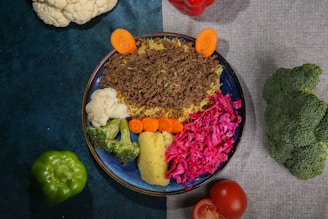 A colorful dish arranged on a blue plate, featuring ground meat over rice, cauliflower, broccoli, carrots, and vibrant red cabbage. Surrounding the plate are whole vegetables including a green bell pepper, tomato, cauliflower, red bell pepper, and broccoli, all placed on a textured surface split between teal and gray.