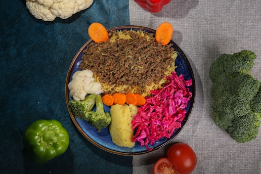 A colorful dish arranged on a blue plate, featuring ground meat over rice, cauliflower, broccoli, carrots, and vibrant red cabbage. Surrounding the plate are whole vegetables including a green bell pepper, tomato, cauliflower, red bell pepper, and broccoli, all placed on a textured surface split between teal and gray.