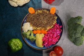 A colorful dish arranged on a blue plate, featuring ground meat over rice, cauliflower, broccoli, carrots, and vibrant red cabbage. Surrounding the plate are whole vegetables including a green bell pepper, tomato, cauliflower, red bell pepper, and broccoli, all placed on a textured surface split between teal and gray.