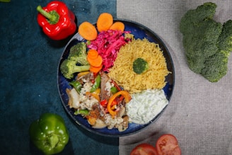 A variety of colorful food items are arranged in a circular blue plate. The plate contains yellow rice topped with a broccoli floret, tzatziki sauce, grilled vegetables and meat, sautéed vegetables including broccoli, red cabbage salad, and sliced carrots. Surrounding the plate are fresh vegetables, including a red bell pepper, green bell pepper, broccoli and two tomato halves, placed on a table partially covered with dark blue and light gray tablecloths.