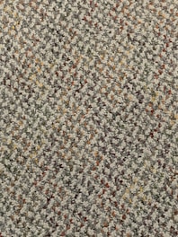 A close-up view of a textured carpet with a speckled pattern. The surface consists of tightly woven loops in various colors, predominantly gray, with interspersed specks of red, orange, yellow, and green.