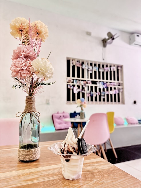 A cozy café interior features a wooden table with a small glass vase holding pink and cream artificial flowers. In the foreground, there is a cup of soft-serve ice cream topped with chocolate syrup and an Oreo cookie. The background shows a seating area with pink and blue cushions and a wall decorated with photos.