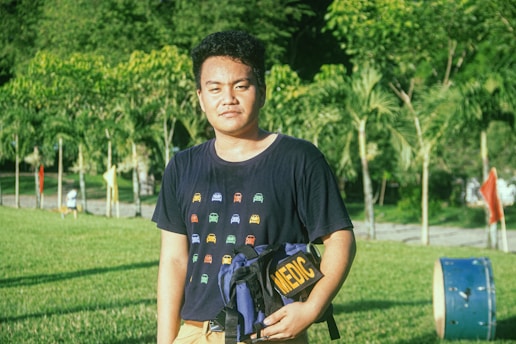 A person stands in a sunlit park holding a dark blue backpack with the word 'MEDIC' on it. They are wearing a dark t-shirt with colorful car illustrations. The background features lush green trees and a clear sky, along with a path lined with small trees and some colorful markers.