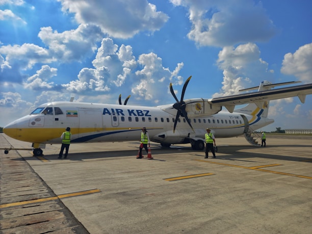 A small commercial propeller aircraft is parked on an airport tarmac. Several people wearing reflective safety vests are standing near the plane, possibly engaged in inspection or maintenance activities. The sky is bright with scattered clouds, creating a clear day setting.