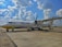 A small commercial propeller aircraft is parked on an airport tarmac. Several people wearing reflective safety vests are standing near the plane, possibly engaged in inspection or maintenance activities. The sky is bright with scattered clouds, creating a clear day setting.