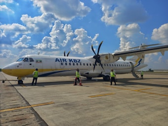A small commercial propeller aircraft is parked on an airport tarmac. Several people wearing reflective safety vests are standing near the plane, possibly engaged in inspection or maintenance activities. The sky is bright with scattered clouds, creating a clear day setting.