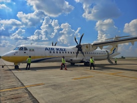 A small commercial propeller aircraft is parked on an airport tarmac. Several people wearing reflective safety vests are standing near the plane, possibly engaged in inspection or maintenance activities. The sky is bright with scattered clouds, creating a clear day setting.