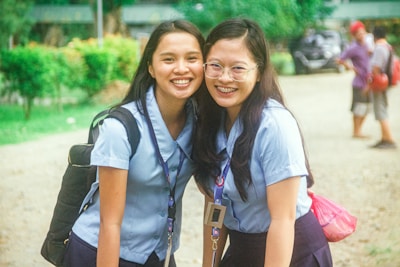 Two young women in matching light blue uniforms are smiling at the camera. One is wearing glasses and has braces, and they both have long dark hair. They are standing outdoors in a green, park-like setting with trees and blurred background elements, including a path on which other people are walking.