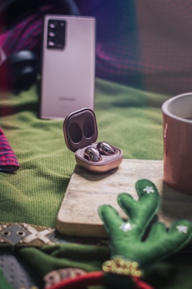 A pair of wireless earbuds in a sleek, metallic charging case is prominently displayed on a wooden board. The background showcases a blurred smartphone with a camera setup, and a mug partially visible on one side. Green fabric and Christmas-themed decorations add festive elements to the scene.