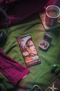 A smartphone displaying a media player screen is placed on a green fabric. Nearby are wireless earphones in an open case, a red and blue plaid shirt, a cup of coffee on a coaster, and a pair of black headphones. A small green decorative object resembling a cactus is visible in the corner.