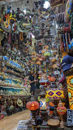 A crowded and colorful shop filled with a variety of antique items and vibrant hanging lamps. The space is densely packed, with shelves and ceilings covered in intricate ornaments, brassware, and handcrafted goods. A person is observed standing in the shop, seemingly engaged in examining or purchasing items.