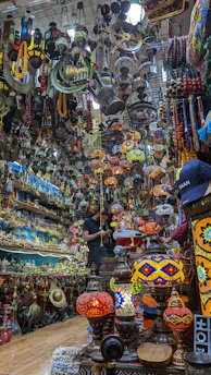 A crowded and colorful shop filled with a variety of antique items and vibrant hanging lamps. The space is densely packed, with shelves and ceilings covered in intricate ornaments, brassware, and handcrafted goods. A person is observed standing in the shop, seemingly engaged in examining or purchasing items.