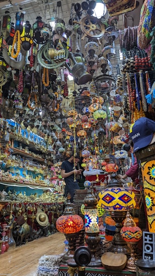 A crowded and colorful shop filled with a variety of antique items and vibrant hanging lamps. The space is densely packed, with shelves and ceilings covered in intricate ornaments, brassware, and handcrafted goods. A person is observed standing in the shop, seemingly engaged in examining or purchasing items.