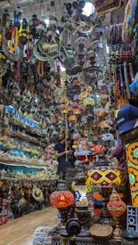 A crowded and colorful shop filled with a variety of antique items and vibrant hanging lamps. The space is densely packed, with shelves and ceilings covered in intricate ornaments, brassware, and handcrafted goods. A person is observed standing in the shop, seemingly engaged in examining or purchasing items.