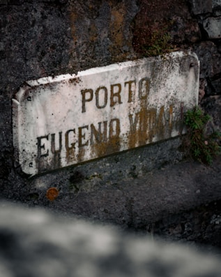 An aged, weathered stone sign with the words 'PORTO EUGENIO VERALLI' is set against a rough stone wall. Moss and small plants are growing around the edges of the sign, adding a natural and rustic feel.