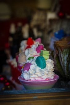 A close-up of a dessert in a glass dish topped with whipped cream and various colorful decorations that resemble small candies or macarons. The background is blurred but contains elements like a tree bark and blurred lights, suggesting a festive or decorative setting.