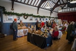 An indoor craft fair is taking place in a hall, decorated with greenery and holiday wreaths on the walls. Various vendors have set up tables displaying handmade crafts, including wooden decorations and woven items. People are browsing and interacting with the vendors, creating a warm and festive atmosphere.