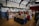 An indoor craft fair is taking place in a hall, decorated with greenery and holiday wreaths on the walls. Various vendors have set up tables displaying handmade crafts, including wooden decorations and woven items. People are browsing and interacting with the vendors, creating a warm and festive atmosphere.