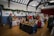 An indoor craft fair is taking place in a hall, decorated with greenery and holiday wreaths on the walls. Various vendors have set up tables displaying handmade crafts, including wooden decorations and woven items. People are browsing and interacting with the vendors, creating a warm and festive atmosphere.