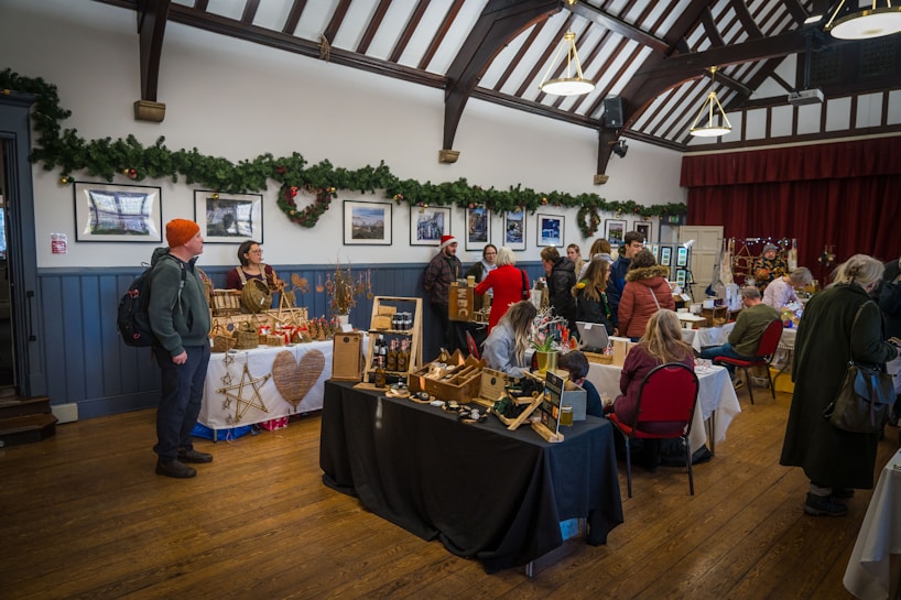 An indoor craft fair is taking place in a hall, decorated with greenery and holiday wreaths on the walls. Various vendors have set up tables displaying handmade crafts, including wooden decorations and woven items. People are browsing and interacting with the vendors, creating a warm and festive atmosphere.