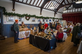 An indoor craft fair is taking place in a hall, decorated with greenery and holiday wreaths on the walls. Various vendors have set up tables displaying handmade crafts, including wooden decorations and woven items. People are browsing and interacting with the vendors, creating a warm and festive atmosphere.