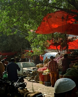 A bustling outdoor market scene with vibrant produce stalls filled with tomatoes, onions, and other vegetables. People are seen moving about, with one person examining the goods. Red cloths provide shade from the sun, adding a colorful backdrop. Motorcycles and a car are parked nearby, while trees provide additional natural cover.