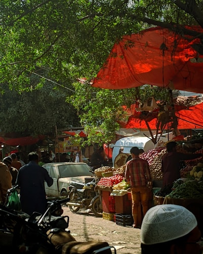 A bustling outdoor market scene with vibrant produce stalls filled with tomatoes, onions, and other vegetables. People are seen moving about, with one person examining the goods. Red cloths provide shade from the sun, adding a colorful backdrop. Motorcycles and a car are parked nearby, while trees provide additional natural cover.
