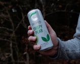 A hand is holding a can labeled 'Celsius Crispy Pear' against a blurred forest background. The can features green and white colors and is marketed as an energy drink with 200 mg of caffeine.