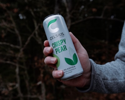 A hand is holding a can labeled 'Celsius Crispy Pear' against a blurred forest background. The can features green and white colors and is marketed as an energy drink with 200 mg of caffeine.