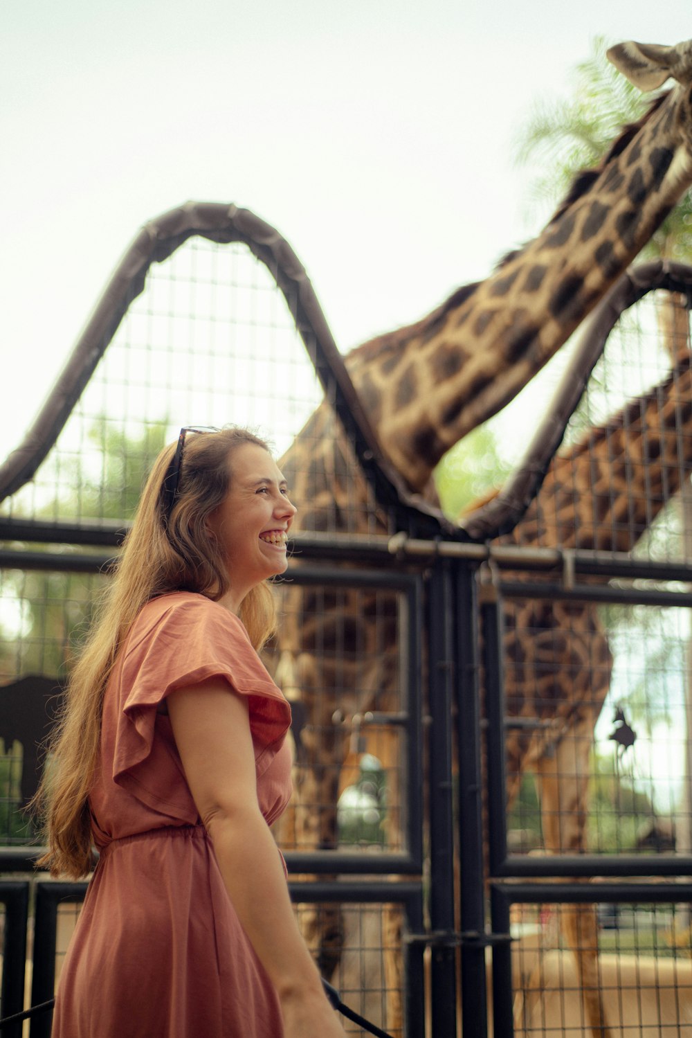 a woman smiling at a giraffe behind a fence