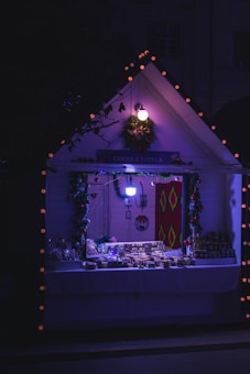 A small, cozy market stall illuminated by soft, warm lights, adorned with Christmas decorations. The stall displays an assortment of handmade goods, possibly desserts or crafts, on a white tablecloth. Wreaths and garlands contribute to a festive atmosphere.