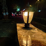 Close-up of a sleek navy outdoor lantern casting warm light on a sandy stone pathway.
