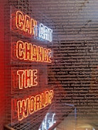 A neon sign with the words 'CAN ART CHANGE THE WORLD?' displayed inside a metal cage. The sign's letters are illuminated in red and white, and the background is filled with numerous names written in a small, uniform font, overlaying across the image.