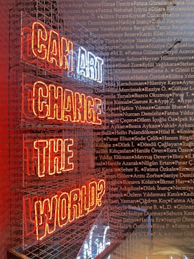 A neon sign with the words 'CAN ART CHANGE THE WORLD?' displayed inside a metal cage. The sign's letters are illuminated in red and white, and the background is filled with numerous names written in a small, uniform font, overlaying across the image.
