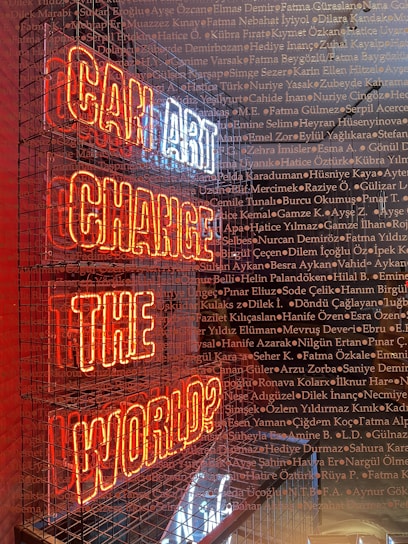 A neon sign with the words 'CAN ART CHANGE THE WORLD?' displayed inside a metal cage. The sign's letters are illuminated in red and white, and the background is filled with numerous names written in a small, uniform font, overlaying across the image.