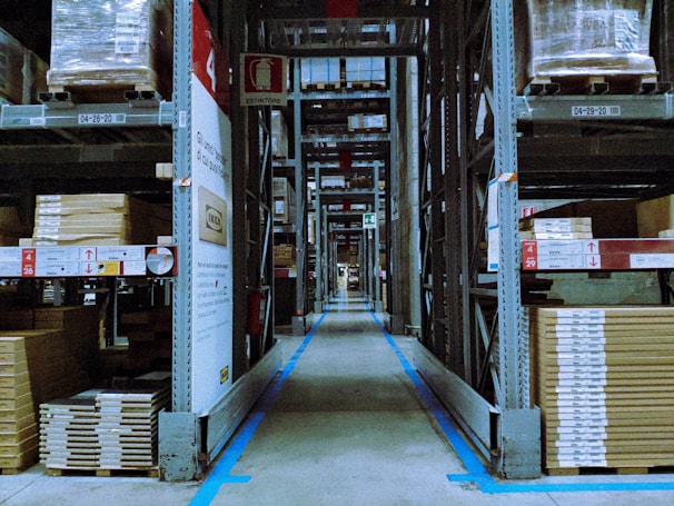A warehouse aisle lined with tall metal shelves stacked with packaged goods. The shelves have labels and signs indicating different sections and product codes. The floor guides with blue lines direct the traffic flow within the facility.